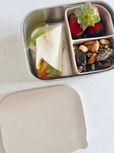 Metal lunch box with compartments containing a wrap, vegetables, and snacks on a white surface.