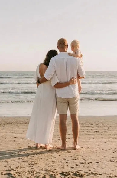 Family of three standing on a beach, looking out at the ocean.