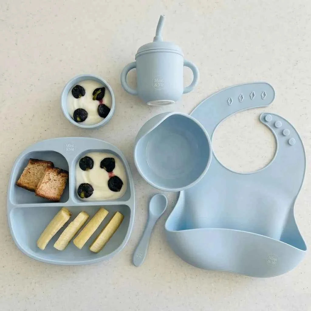 Baby mealtime set with blue tray, bowl, cup, bib, and spoon on a light background